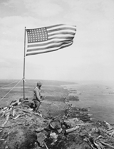 From the crest of Mount Suribachi, the Stars and Stripes wave in triumph over Iwo Jima after U.S. Marines had fought their way inch by inch up its steep lava-encrusted slopes. ca. 02/1945. Photo Credit: National Archives 